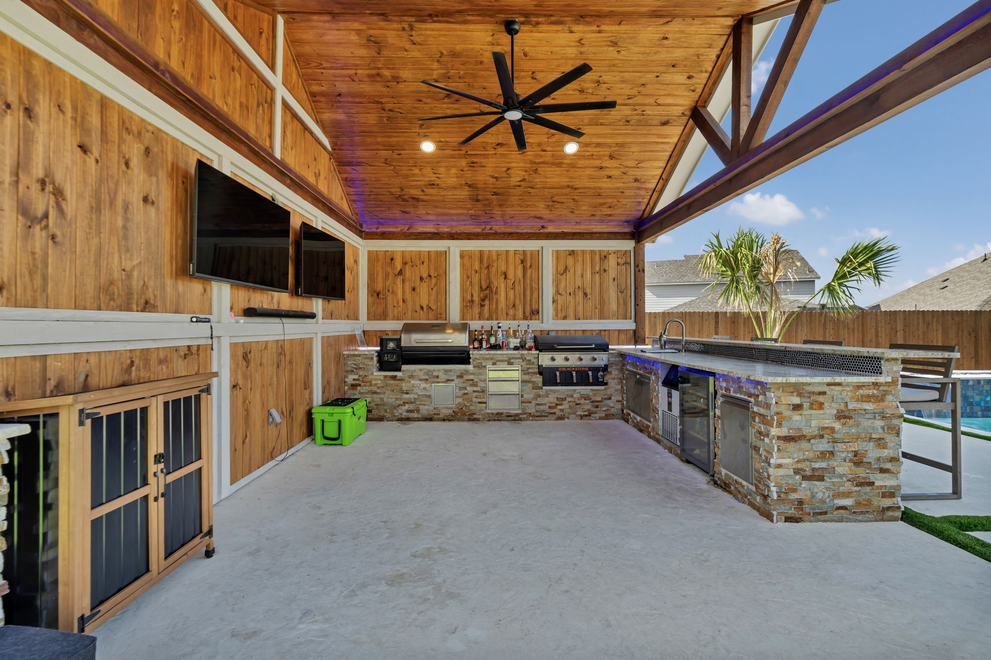 Outdoor kitchen under a timber pavilion with a ceiling fan and TVs